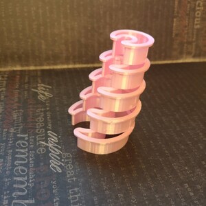 May include: A light pink, 3D-printed spiral staircase structure composed of five stacked, crescent-shaped tiers. The object features a smooth, uniform finish and a vertical, curved design that tapers slightly toward the top, creating a tiered effect.