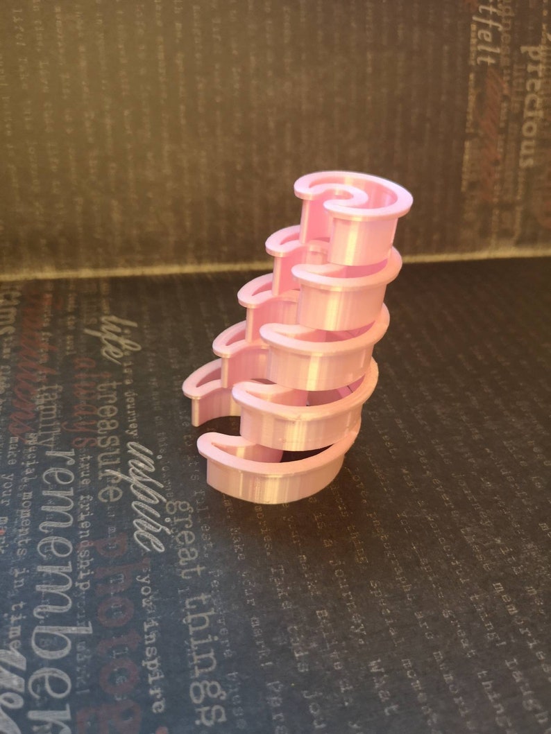 May include: A light pink, 3D-printed spiral staircase structure composed of five stacked, crescent-shaped tiers. The object features a smooth, uniform finish and a vertical, curved design that tapers slightly toward the top, creating a tiered effect.