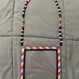 May include: A clear, square-shaped accessory with a rainbow-colored border and a black frame. The border is made of small, colorful beads. The strap is made of black and white beads, and rainbow beads.