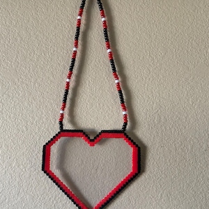 May include: A beaded heart-shaped decoration with a clear center. The heart is outlined in red and black beads, with a beaded strap in black, red, and white. The decoration is hanging against a beige wall.