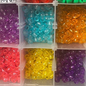 May include: A clear plastic organiser filled with colourful beads. The beads are in various colours, including purple, blue, orange, red, yellow, and white. The beads are small and cylindrical, ideal for crafting and jewellery making.