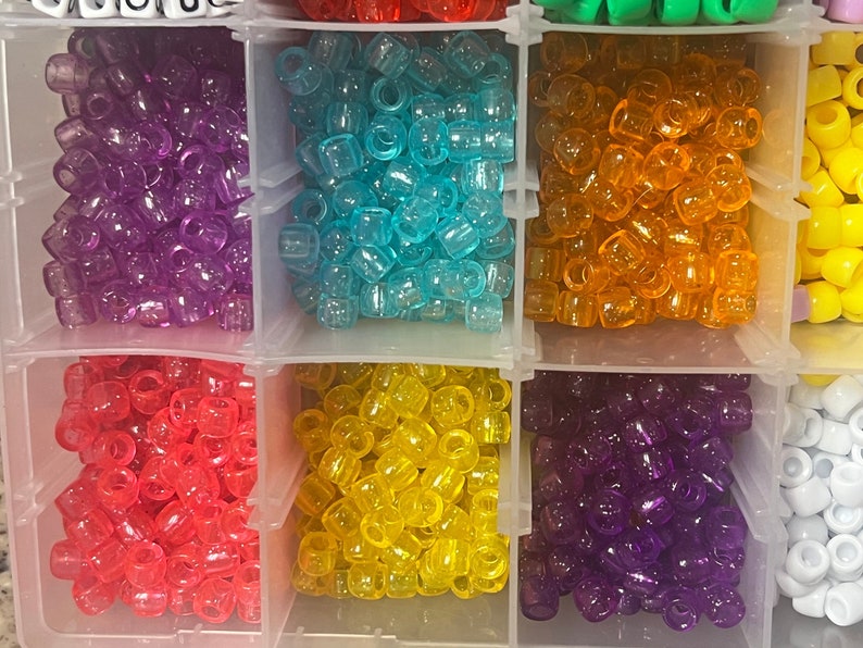 May include: A clear plastic organiser filled with colourful beads. The beads are in various colours, including purple, blue, orange, red, yellow, and white. The beads are small and cylindrical, ideal for crafting and jewellery making.