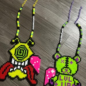 May include: Two beaded necklaces with pendant designs. One features a red, yellow, and white monster with a pink wing. The other is a green and purple teddy bear with the text "LUL UP" and a pink wing. Both have beaded chains.