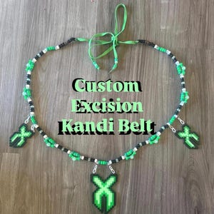 May include: A beaded belt featuring a pattern of black, white, and green pony beads with three green and black X-shaped charms hanging from silver-toned metal rings. The belt includes green fabric ties at the top and displays the text Custom Excision Kandi Belt.