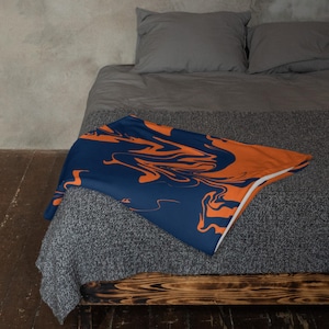 May include: A navy blue and orange throw blanket with a marbled design. The blanket is folded over a bed, showcasing the contrasting colours and abstract pattern. The blanket's edges are white.