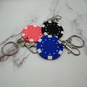 May include: Three circular keychains shaped like casino poker chips in pink, black, and blue. Each chip features a white rectangular pattern along the outer edge and is attached to a silver-toned metal split ring and a lobster claw clasp.
