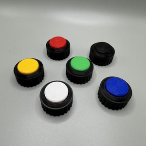 May include: A collection of six circular push buttons, each featuring a black ridged base and a flat, colored top. The set includes individual buttons with red, yellow, green, white, blue, and black circular tops arranged in a scattered formation.