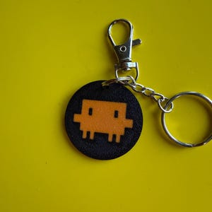 May include: A circular black keychain featuring a pixelated orange character design. The keychain is attached to a silver-colored metal lobster clasp and a circular split ring, providing a secure way to attach the item to keys or bags.