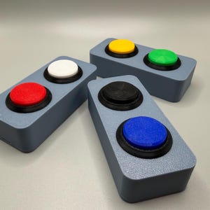 May include: Three rectangular gray plastic devices, each featuring two circular push buttons. The buttons are colored red, white, yellow, green, black, and blue. Each button is set within a black circular frame mounted on the gray base.