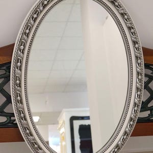 Op de afbeelding: Ovale spiegel met een witte lijst en sierlijke zilveren details. De lijst van de spiegel heeft een decoratief patroon en een kralen binnenrand. De spiegel is op een houten oppervlak gemonteerd.