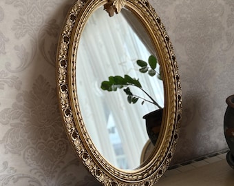 May include: Oval gold-framed mirror with ornate detailing. The mirror reflects a room with a plant and curtains. A small gold mask and a black and white electronic device are on the table.