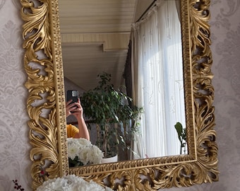 May include: A rectangular gold-colored mirror with an ornate, carved frame. The frame features a detailed, scrolling leaf design. The mirror reflects a room with white curtains, a plant, and a person taking a photo.
