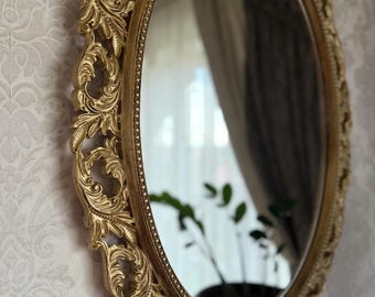 May include: An oval wall mirror featuring an ornate gold-colored frame with intricate scrollwork and leaf-like carvings. The frame has a beaded inner border surrounding the reflective glass surface, which shows a reflection of a plant and curtains.