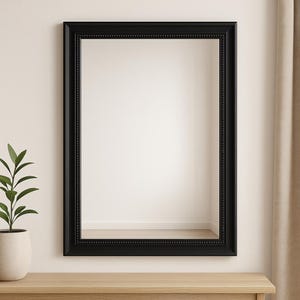 Puede incluir: Un espejo rectangular con marco negro y borde interior con cuentas. El espejo está montado sobre una superficie de madera clara, con una planta en maceta a la izquierda. El marco tiene un diseño clásico y decorativo.