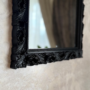 Op de afbeelding: Een rechthoekige zwarte spiegel met een sierlijk, gesneden frame. Het frame heeft gedetailleerd krulwerk en een kralen binnenrand. De spiegel reflecteert een kamer met een neutraal getinte muur en een gordijn.