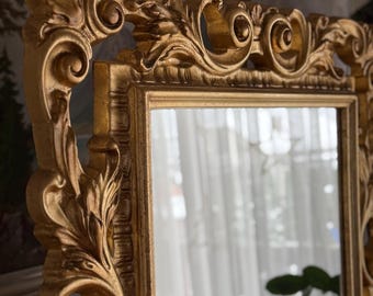 May include: A gold-colored, ornate mirror with a detailed, carved frame. The frame features scrolling leaf and floral designs. The mirror reflects a blurred view of a window and a plant. The overall style is classic and elegant.