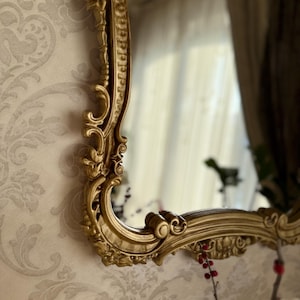 Może przedstawiać: Ozdobne, złote lustro z bogato zdobioną ramą. Lustro jest zamontowane na ścianie z jasną tapetą w wzory. Rama ma klasyczny, zabytkowy design.