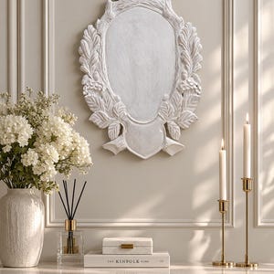 Puede incluir: Una placa de pared ovalada de color blanco con detalles tallados de hojas de roble y bellotas, rematada con un motivo de cinta. La placa tiene un acabado desgastado y está montada en una pared sobre una superficie con flores.