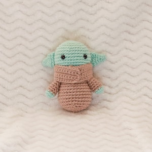 May include: A crocheted plush toy in the shape of a green alien with large ears and a pink crocheted coat. The toy has black eyes and a small mouth.