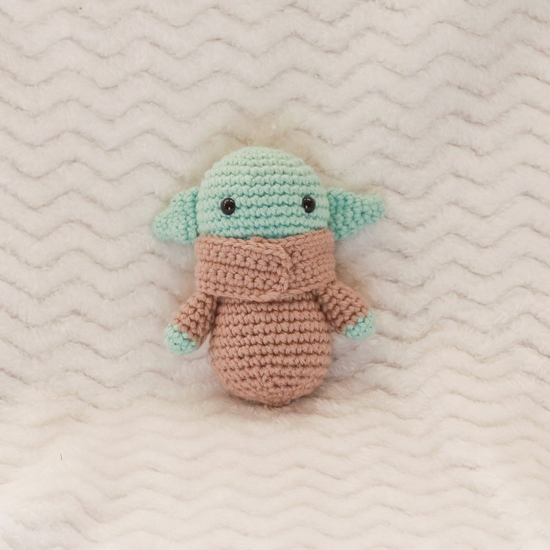 May include: A crocheted plush toy in the shape of a green alien with large ears and a pink crocheted coat. The toy has black eyes and a small mouth.