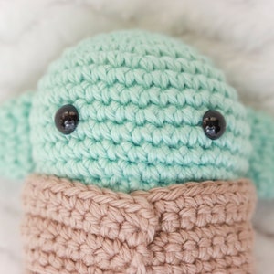 May include: A crocheted green and brown plush toy with black eyes. The toy is shaped like a small alien with large ears.