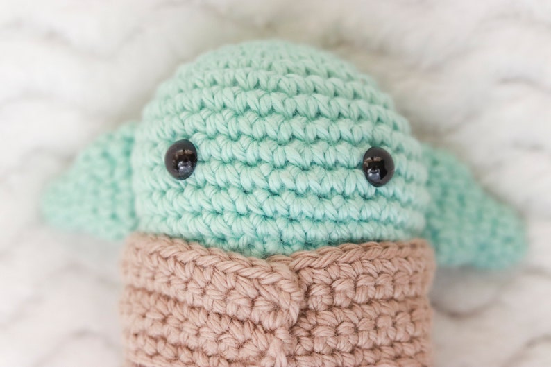 May include: A crocheted green and brown plush toy with black eyes. The toy is shaped like a small alien with large ears.