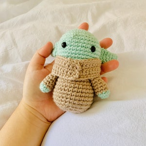 May include: A crocheted green and brown plush toy resembling a character from the Star Wars franchise. The toy has large black eyes and a small mouth.