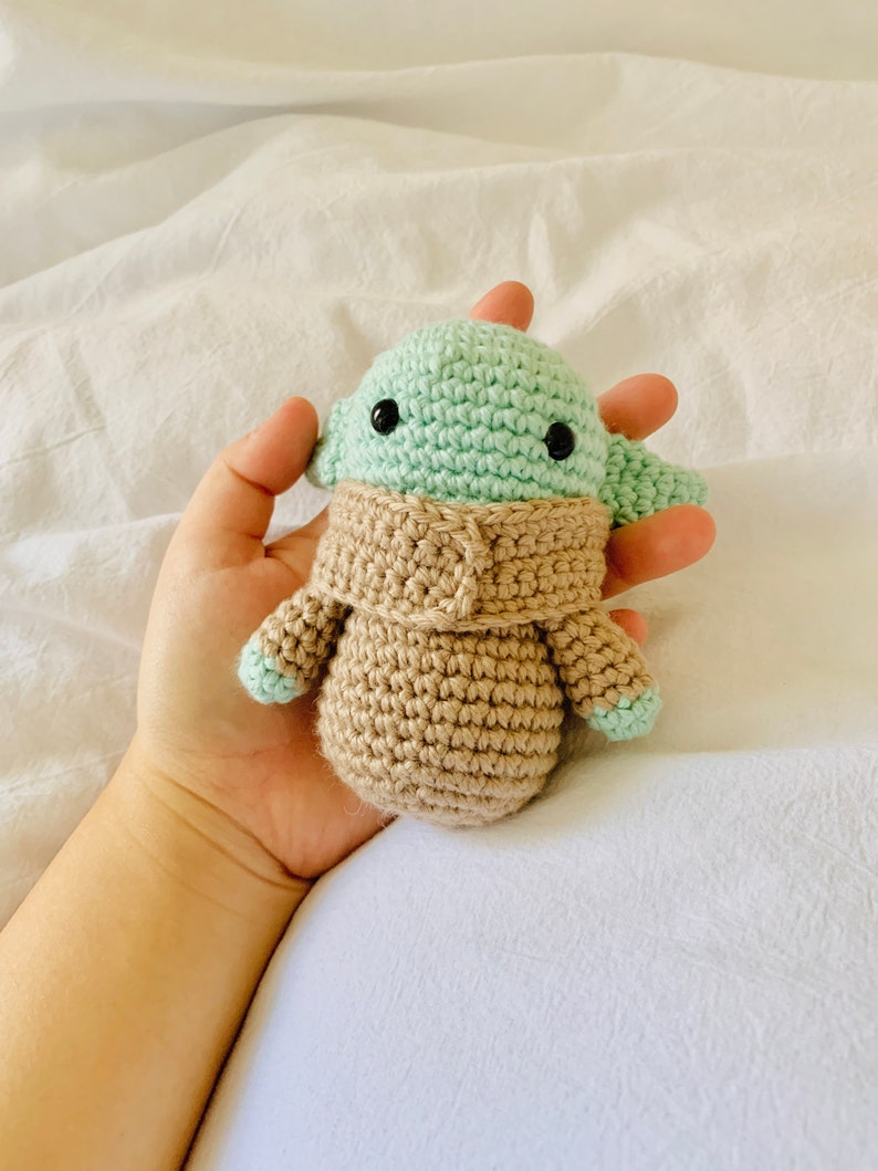 May include: A crocheted green and brown plush toy resembling a character from the Star Wars franchise. The toy has large black eyes and a small mouth.