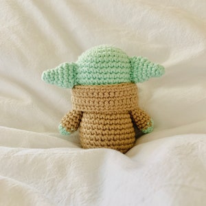 May include: A crocheted plush toy of a popular character from the Star Wars franchise. The toy is green and brown and has large ears.