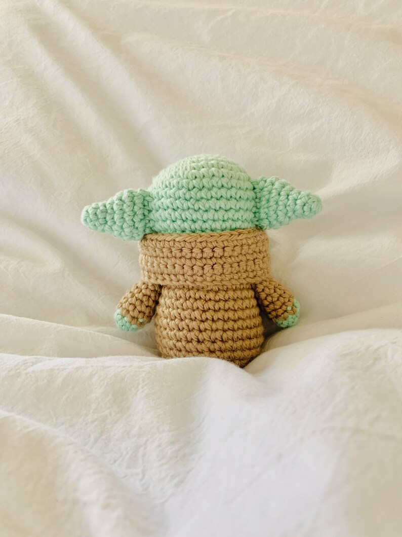 May include: A crocheted plush toy of a popular character from the Star Wars franchise. The toy is green and brown and has large ears.