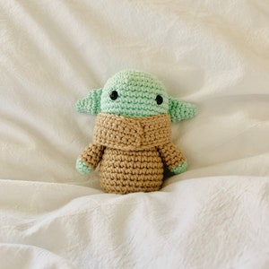 May include: A crocheted plush toy of a green alien character with large eyes and a brown robe. The toy is sitting on a white fabric surface.