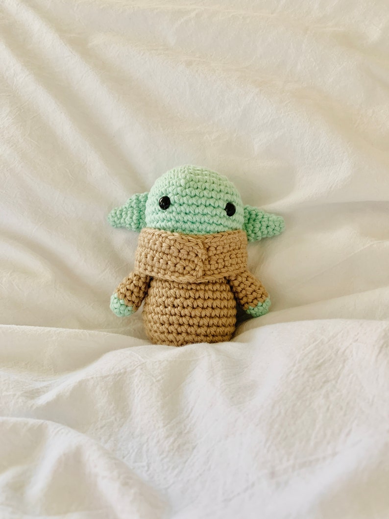 May include: A crocheted plush toy of a green alien character with large eyes and a brown robe. The toy is sitting on a white fabric surface.