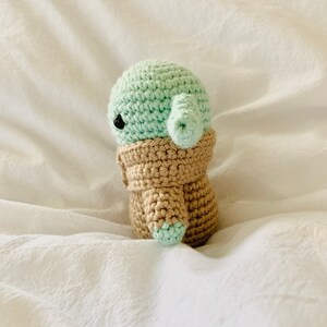 May include: A crocheted plush toy of a green character with a brown coat. The toy is sitting on a white surface.
