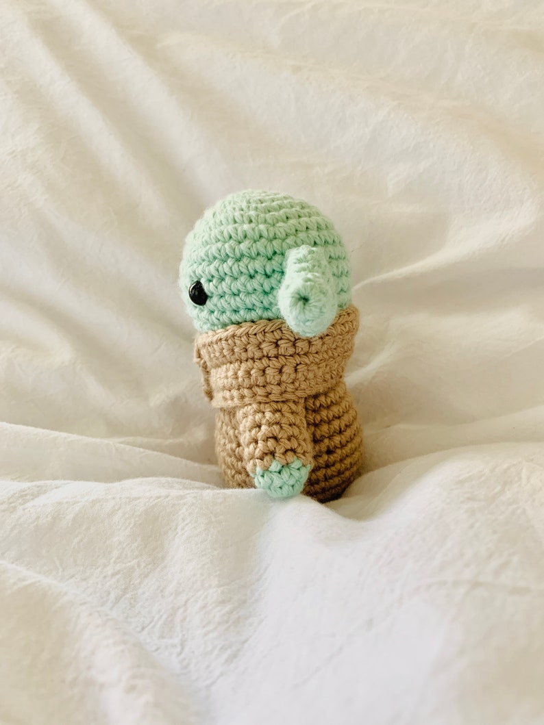 May include: A crocheted plush toy of a green character with a brown coat. The toy is sitting on a white surface.