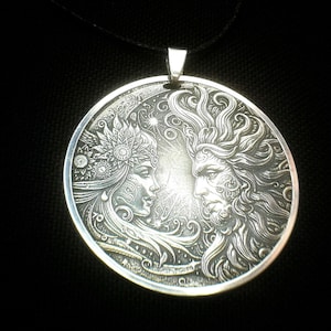 Può includere: Una collana con pendente in argento con un rilievo dettagliato di due volti, uno ornato da un copricapo floreale, l'altro con capelli e barba fluenti. Il pendente rotondo è sospeso a un cordino nero.