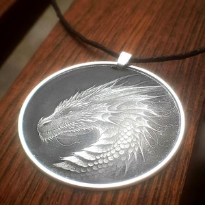 以下が含まれることがあります： 詳細なドラゴンのデザインが施されたシルバーペンダント。ドラゴンの頭部は、円形のフレーム内の黒い背景に配置されています。ペンダントは黒いコードから吊り下げられており、ファンタジーをテーマにしたアクセサリーです。