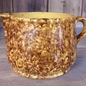 May include: A speckled brown and yellow ceramic pitcher with a handle and a spout. The pitcher has a rounded body and a slightly flared rim. The handle is arched and attached to the side of the pitcher. The pitcher is likely vintage or antique.
