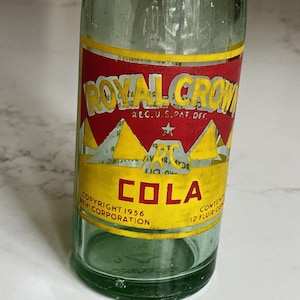 May include: Vintage green glass Royal Crown Cola bottle. The label features the words "ROYAL CROWN COLA" in yellow and red, with the text "COPYRIGHT 1936 NEW CORPORATION" and "12 FLUID OZ".
