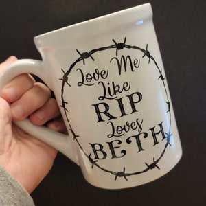 May include: White ceramic mug with a black design of barbed wire around the words "Love Me Like RIP Loves Beth".