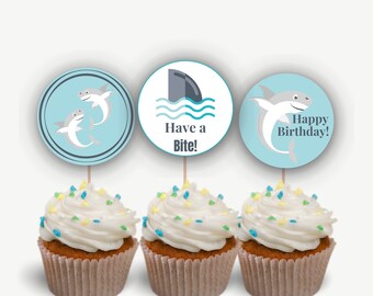 May include: Three cupcake toppers with blue and white shark designs. The toppers say "Have a Bite!" and "Happy Birthday!"
