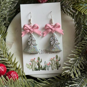 May include: A pair of dangle earrings featuring a silver-toned glittery Christmas tree shape topped with a light pink bow. Each tree is decorated with a vertical row of clear marquise-cut crystals. The earrings are displayed on a white card.