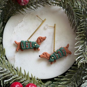 Peut inclure: Une paire de boucles d'oreilles pendantes représentant des teckels marron vêtus de pulls verts à pois rouges avec une petite étoile dorée. Les chiens sont fixés à de fines tiges métalliques dorées avec des fermoirs en plastique transparent.