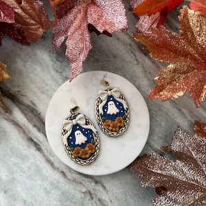 Peut inclure: Une paire de boucles d'oreilles pendantes ovales avec un fond bleu foncé, un fantôme blanc, des petits points blancs et des fleurs orange. Chaque boucle est encadrée par une bordure dorée festonnée et surmontée d'un nœud blanc, avec des crochets dorés.