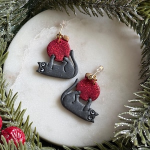 May include: A pair of drop earrings featuring a dark grey cat figure holding a round, glittery red bauble. The cat has white eyes with black pupils, and the bauble is attached to a gold-toned metal post with a clear plastic backing.