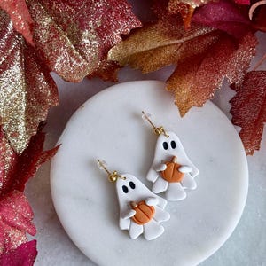 May include: A pair of white ghost-shaped earrings with black eyes and a small orange pumpkin held in the centre. Each earring features a gold-toned post backing and a smooth finish, showcasing a playful design that captures the essence of autumn.