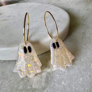 May include: A pair of hoop earrings featuring ghost-shaped charms. The charms are translucent white with iridescent glitter flakes embedded throughout and have two small, solid black oval shapes representing eyes on each ghost figure.