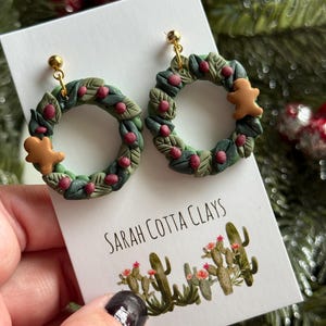 Peut inclure: Une paire de boucles d'oreilles pendantes en forme de couronnes de Noël, avec des feuilles vertes, de petites baies rouges et un bonhomme en pain d'épice marron sur chacune. Elles sont fixées sur un carton blanc portant le texte SARAH COTTA CLAYS.