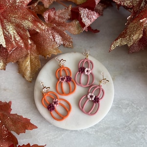 May include: Two pairs of dangle earrings shaped like pumpkins. One pair is bright orange and the other is dusty rose. Each earring features a small cluster of tiny dark red and light pink flowers at the centre and a clear crystal stud top.