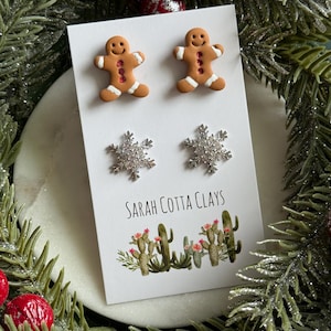 May include: A set of two pairs of stud earrings on a white display card. The top pair features brown gingerbread men with white icing details and red accents. The bottom pair consists of silver-toned snowflake studs with clear crystals.