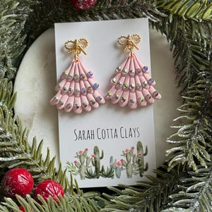 Peut inclure: Une paire de boucles d'oreilles rose clair en forme de sapin à étages, ornées de nœuds dorés sur le dessus et de strass irisés. Elles sont présentées sur une carte blanche marquée Sarah Cotta Clays avec une illustration de cactus.